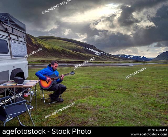 Man playing guitar while relaxing by off-road vehicle against cloudy sky during sunset