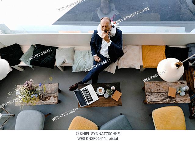 Businessman sitting relaxed in coffee shop with unicorn pillow around his neck, overhead view