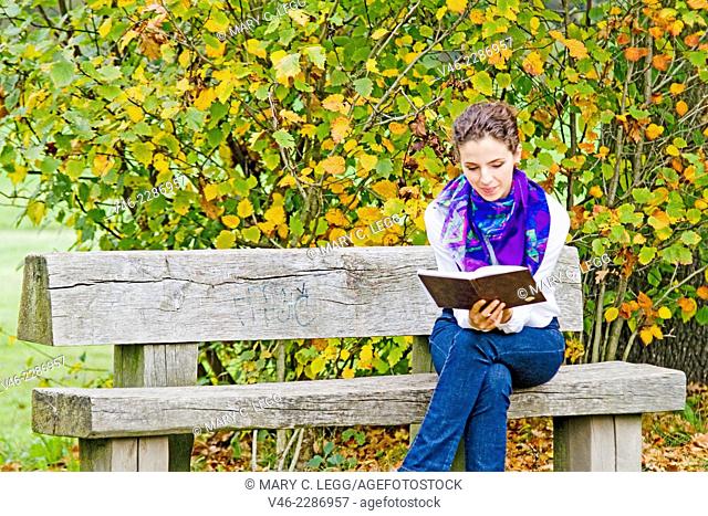 Woman on a park bench with book. Woman in white blouse with blue scarf and blue jeans is sitting on bench in a park