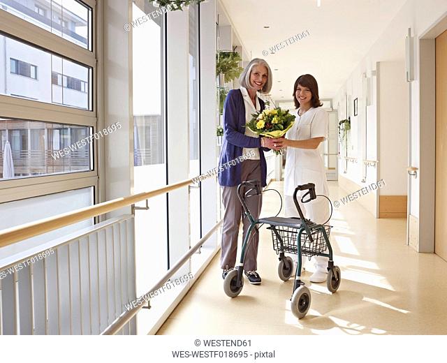 Germany, Cologne, Senior women and caretaker holding bouquet and standing by wheeled walker, smiling