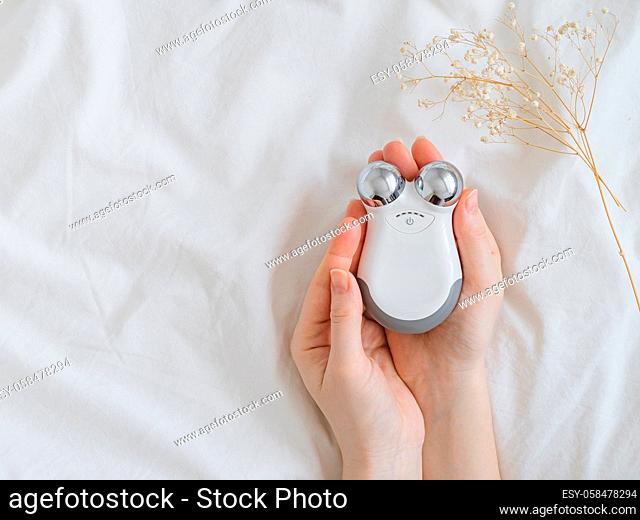 Microcurrent concept. Face massager with micro currents for home use and skincare on white bed. Female hands holds device for anti-aging and face lifting