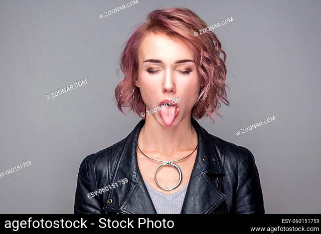 Closeup portrait of funny closed eyes beautiful girl with short hairstyle and makeup in casual style black leather jacket standing with tongue out