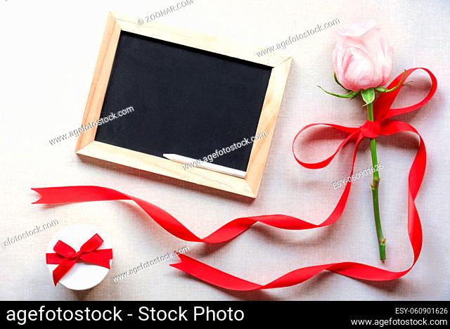 Pink rose tied with red ribbon and bow, near a cute white gift box and a blank chalkboard, on a vintage fabric background