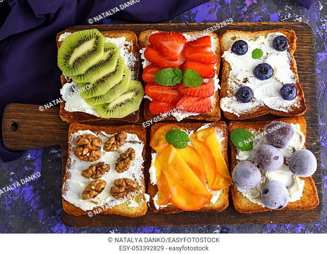 French toasts with soft cheese, strawberries, kiwi, walnuts, cherries and blueberries on a brown wooden board, top view