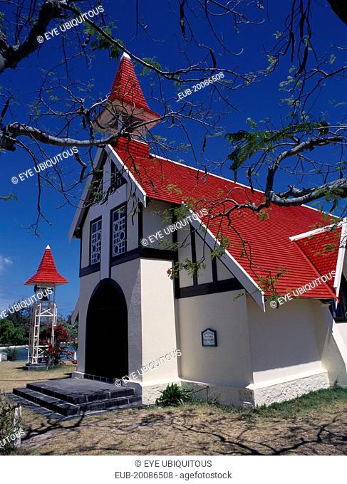 Marie-Reine Church. White painted exterior with red tiled roof and free standing bell tower, part framed by tree branches