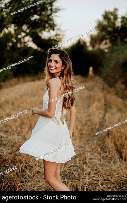 Smiling beautiful woman wearing white dress standing in wheat field during sunset