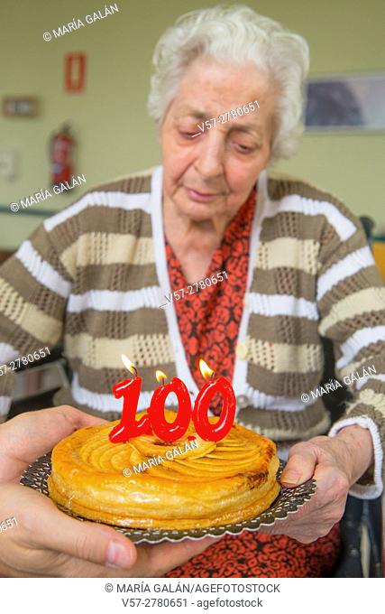 Old woman in a nursing home, on her one hundred birthday, blowing birthday's candles on a cake held by a man's hands