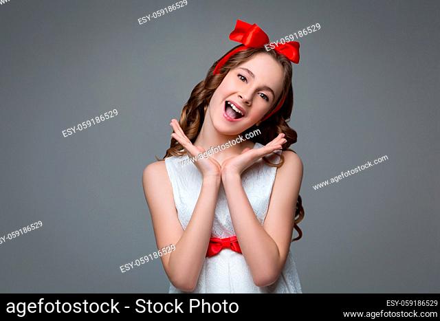 Beautiful teenage girl with long curly hair and red ribbon bow on head wearing white dress. Happy surprised expression. Studio portrait on grey background