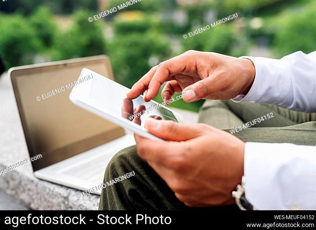 Male professional with laptop using digital tablet while sitting on retaining wall