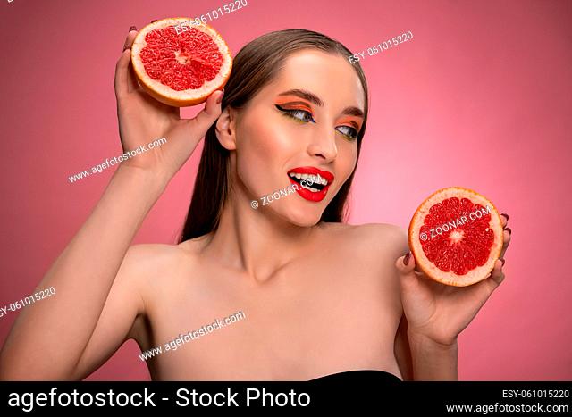 Beautiful young woman model holding two juicy grapefruit sliced in half in her hands looking sideways. Charming joyful funny lady with red lips and long hair...
