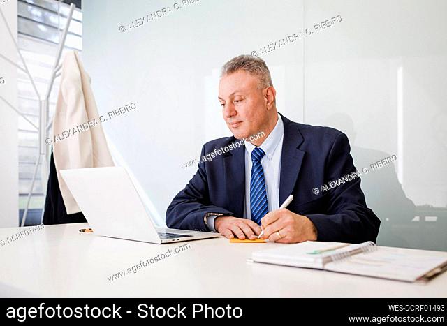 Mature businessman using laptop writing in sticky note at desk in office