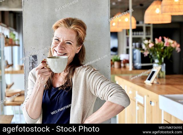 Happy mature woman with hand on hip having coffee against column at home