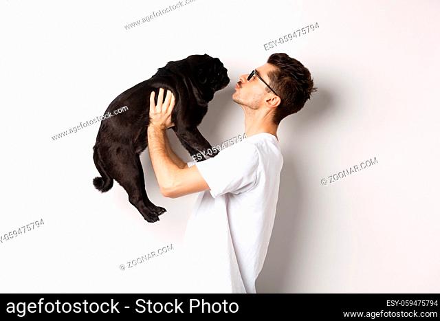 Profile of handsome young man kissing small cute dog face. Hipster guy loving his pug, standing over white background