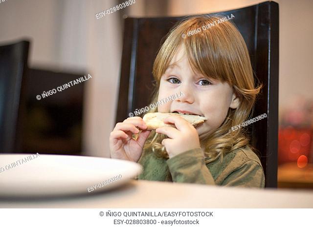 Three years old blonde hungry child biting pizza piece in her hands, sitting in dark brown chair, in table with grey tablecloth at home, looking at camera