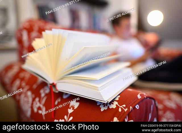 PRODUCTION - 18 April 2023, Saxony, Dresden: ILLUSTRATION - An open book lies on a sofa back, while a woman sits on the sofa holding a book in her hands