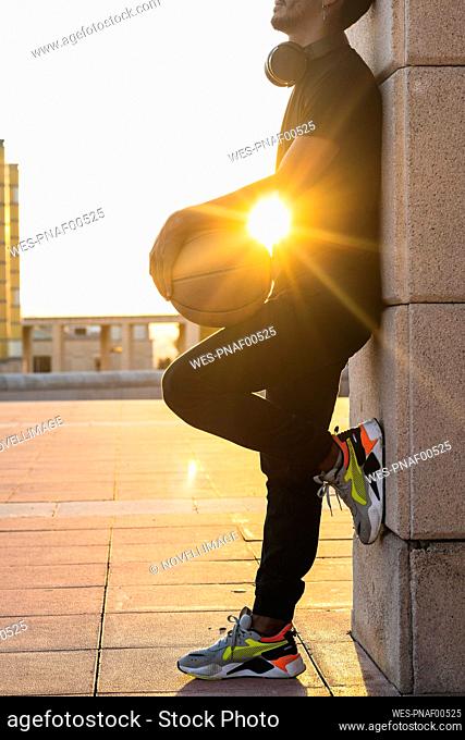 Sun shining through young man holding basketball while standing by wall at sunset