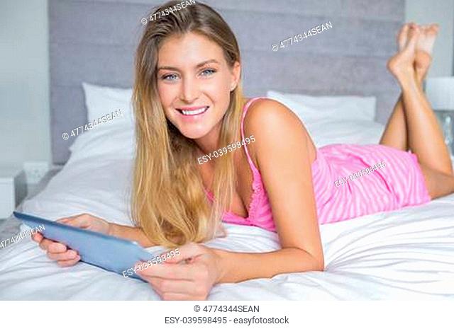 Happy blonde lying on her bed using her digital tablet smiling at camera in bedroom at home