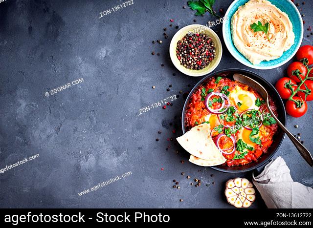 Shakshuka with pita bread in pan, hummus in bowl on rustic background. Middle eastern traditional dishes. Fried eggs with vegetables. Top view
