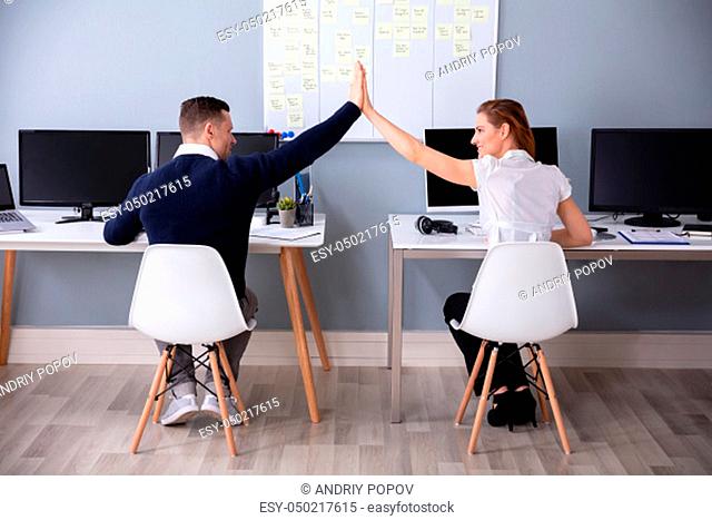 Rear View Of Businesspeople Sitting On Chair Giving High Five In Office