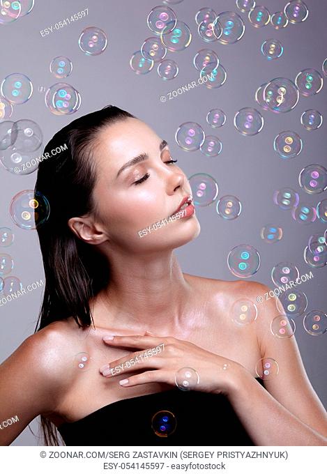 Beautiful young brunette woman with soap bubbles on grey background. Female with eyes closed