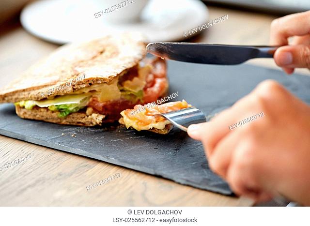food, dinner and people concept - hand of person eating salmon panini sandwich with tomatoes and cheese using fork and knife at restaurant