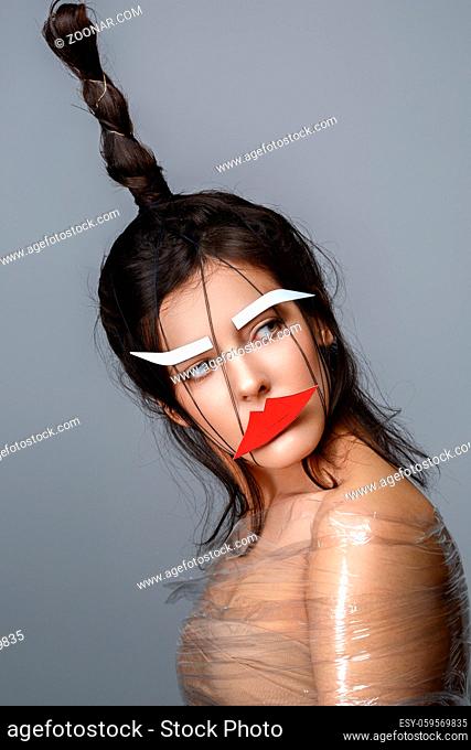 Beautiful young woman with odd fancy hairstyle, body wrapped in plastic foil and eyebrows and lips paper cutouts on face