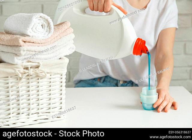 Close up of female hands pouring liquid laundry detergent into cap on white rustic table with towels on background in bathroom