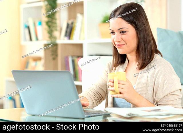 Satisfied woman drinking coffee using a laptop on a desk at home