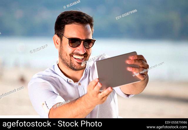 Handsome man with sunglasses using tablet for web chatting on beach. Enjoying beside river