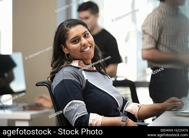 Young woman sitting at computer in office looking to the camera smiling, co-workers having discussion around dry erase board in background