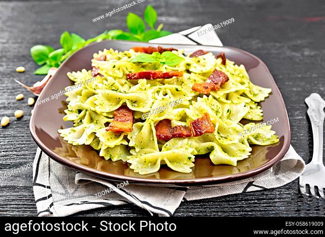Farfalle pasta with pesto sauce, fried bacon and basil in a plate on a napkin on black wooden board background