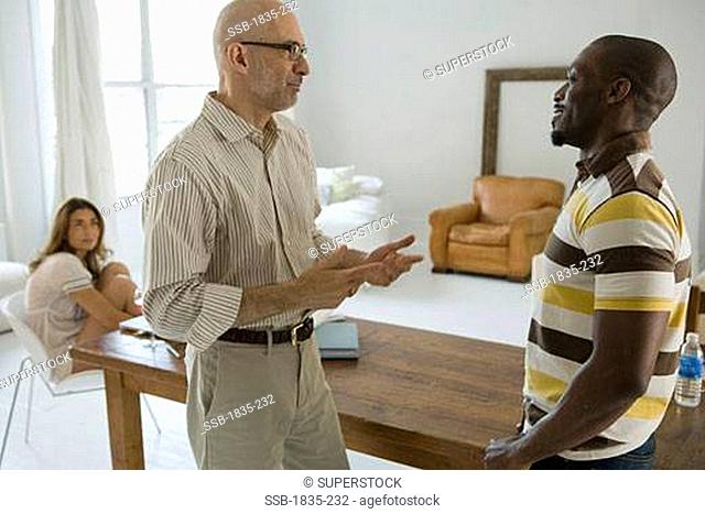 Mature man talking to a mid adult man with a woman sitting on a chair in the background