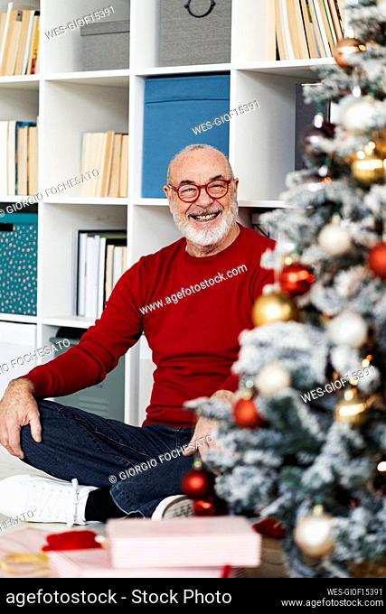 Happy senior man looking at Christmas tree sitting in front of bookshelf