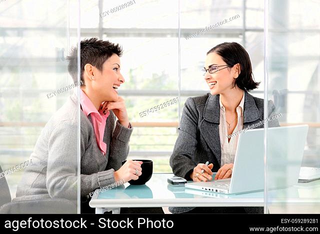 Happy young businesswomen working together at desk in modern office, looking at each other, laughing