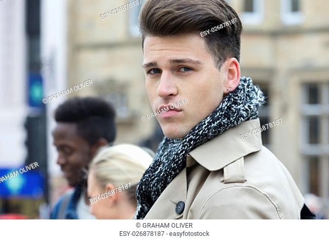 A young male looks at the camera as he stands outside in the city centre with friends. He is wrapped up in a coat and scarf for the cold weather