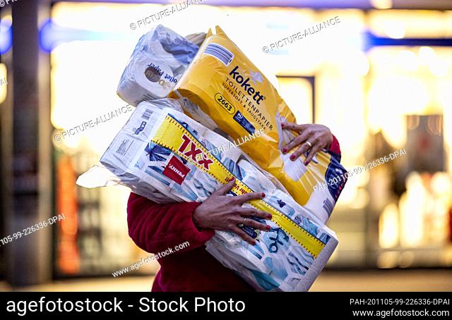 05 November 2020, Hamburg: A woman walks out of a drugstore with an arm full of rolls of toilet paper. Yawning empty shelves where the toilet paper used to sit...
