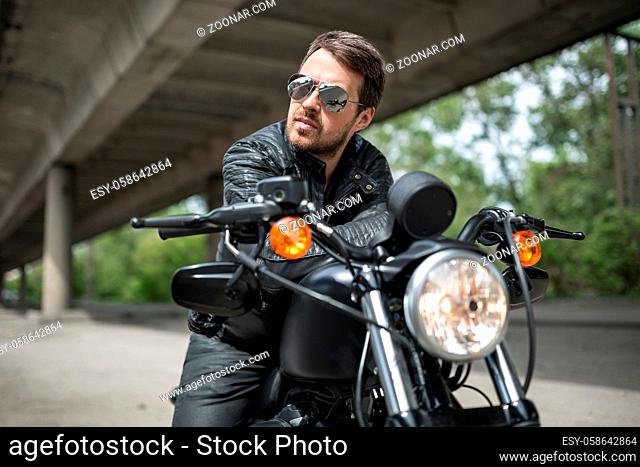 Charismatic man on the black motorbike on the background of the overpass and trees. He holds his hands on the motorbike. He looks to the right