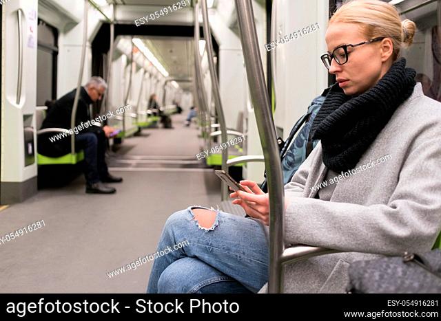 Beautiful blonde caucasian woman wearing winter coat and scarf reading on the phone while traveling by metro. Public transport