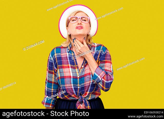 Throat pain illness or flu. Portrait of sick modern stylish mature woman in casual style with hat and eyeglasses standing and holding her painful neck