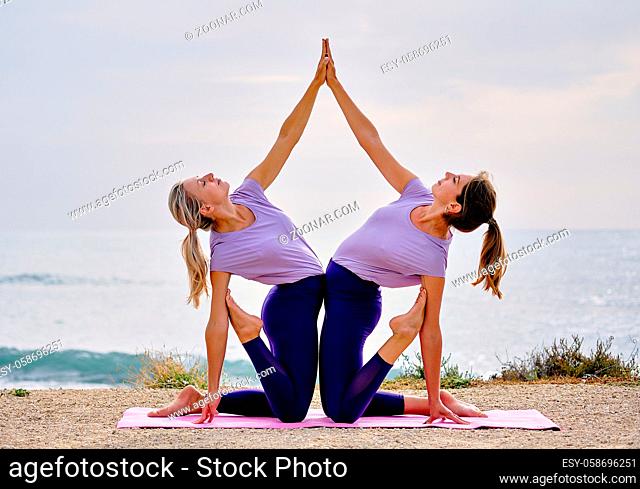 Two slim european sportswomen wearing equal the same activewear doing acro tandem yoga on mat on coast near the sea in the morning