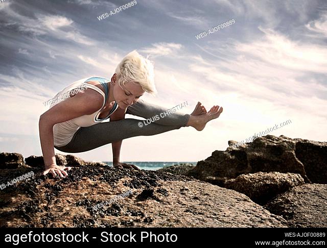 Blond woman practicing Astavakrasana on rock formation at beach against sky