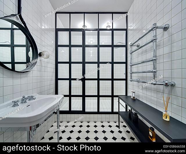Bathroom with walls of white tiles. There is white washbasin, black mirror, shower with black partition of glass squares and glowing lamps