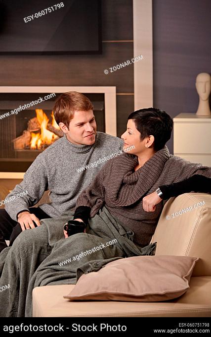 Young couple hugging on sofa in front of fireplace at home, looking at each other, smiling