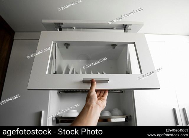 Hand holds the door of the dryer with utensils. Dish drying metal rack with white clean plates. Traditional comfortable kitchen