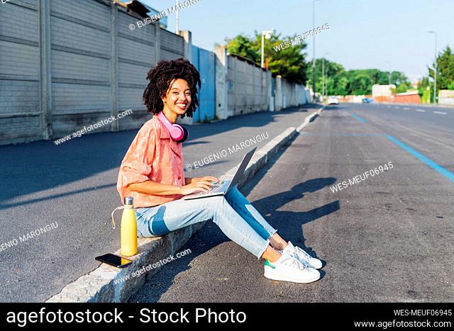 Happy young woman wearing wireless headphones sitting with laptop on road