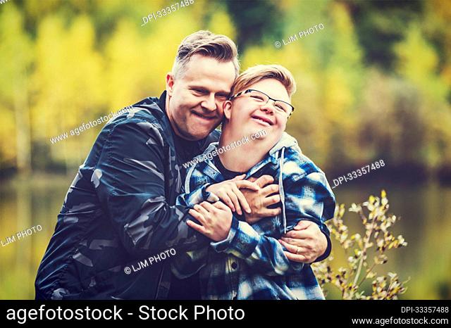 A young man with Down Syndrome and his father enjoying each other's company and giving each other a hug after pretending to wrestle in a city park on a warm...