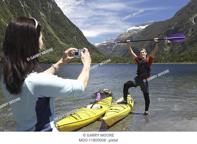 Woman taking picture of man hoisting oar of kayak over head on shore of mountain lake