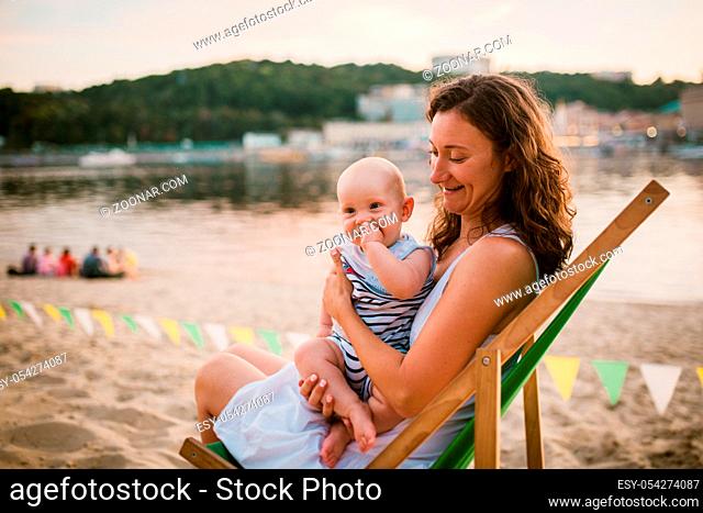 Young Caucasian woman, beautiful mother holds the child in her arms, mom hugs her one-year-old son sitting in a chair on the sandy beach in the park