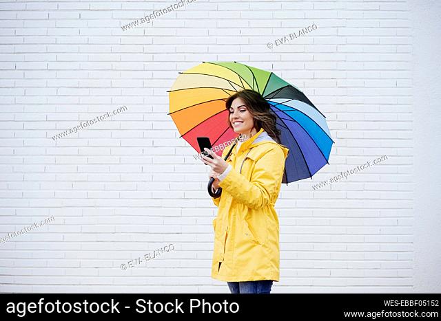 Woman with umbrella using mobile phone in front of wall