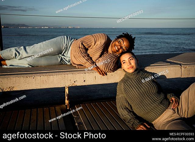 Young woman looking away while sitting by friend lying on bench against sea